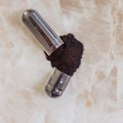 Närbild på en öppen kapsel liggandes på marmor med mörkt pulver som runnit ut